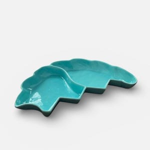 May include: A turquoise ceramic serving dish with a unique, wave-like design. The dish is divided into two sections and has a glossy finish. The overall shape is reminiscent of a stylized cloud or abstract form, perfect for serving appetizers or snacks.
