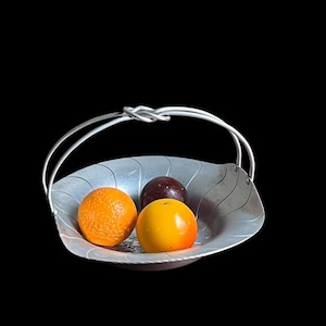 May include: A silver-colored metal basket with a handle, holding three pieces of fruit. The fruit includes an orange, a yellow-orange fruit, and a dark purple fruit. The basket has a leaf-like design.