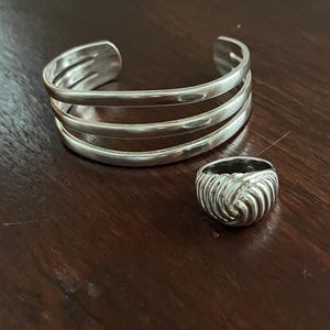 May include: A silver cuff bracelet with three parallel bands and a matching silver ring with a textured, knotted design. The jewelry is displayed on a dark brown wooden surface.