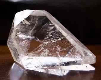May include: A large, clear quartz crystal with a pointed tip and many facets. The crystal is resting on a dark brown surface.