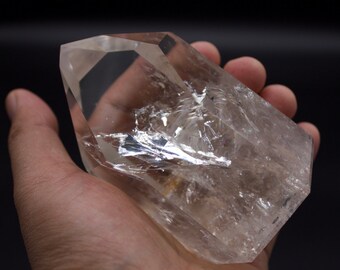 May include: A large, clear quartz crystal with a point on one end. The crystal is clear and has a slight shimmer to it. The crystal is being held in a hand.