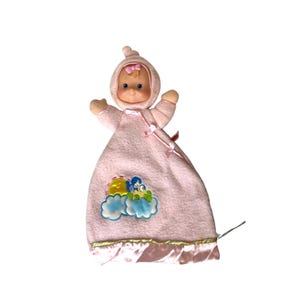 May include: A vintage baby doll with a pink fleece blanket. The doll has a pink hood with a bow, and a smiling face. The blanket features a cloud and flower applique and a satin trim.