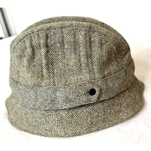 May include: A herringbone patterned hat in shades of gray, tan, and black. The hat features a rounded crown, a structured brim, and a band around the base with a single black button detail. The hat is a classic style.