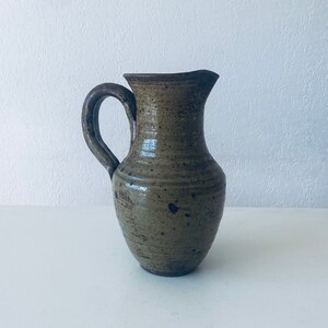 Puede incluir: Una jarra de cerámica marrón con un esmalte moteado. La jarra tiene un cuerpo redondeado y un asa.