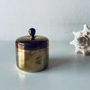 May include: A small, round, brass container with a lid. The container has a smooth, polished surface and a slightly domed lid with a small knob on top. The container is sitting on a white surface.