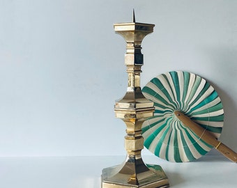 May include: A tall, brass candlestick holder with a square base and a spike at the top. A decorative fan with green and white stripes is behind the candlestick. The fan has a wooden handle.