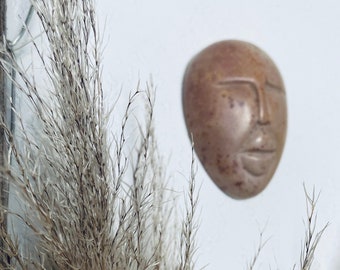 Könnte beinhalten: Ein braunes, ovales Wandobjekt mit einem geschnitzten Gesicht, das an einer weißen Wand befestigt ist. Das Gesicht hat geschlossene Augen und einen ruhigen Ausdruck. Getrocknetes, beiges Ziergras befindet sich im Vordergrund.
