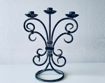 Puede incluir: Un candelero de metal negro con tres brazos y un diseño decorativo de volutas. El candelero tiene una base circular y está en un acabado desgastado.
