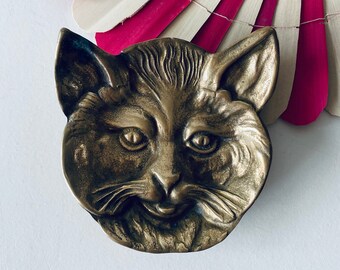 Puede incluir: Un plato de metal para joyas con forma de cara de gato. El plato tiene un diseño detallado con una superficie texturizada y una representación realista de los rasgos de un gato.