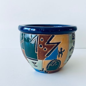 Peut inclure: Un petit pot rond en céramique avec un bord bleu. Le pot est décoré d'un motif géométrique dans des tons de brun, vert, bleu et jaune.