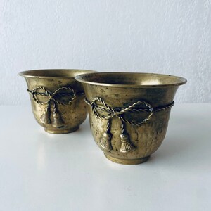 Peut inclure: Deux petits pots en laiton avec un design en corde et des glands. Les pots sont sur une surface blanche.