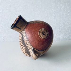 Puede incluir: Una olla de cerámica marrón y roja con diseños pintados en negro y blanco. La olla tiene un cuerpo redondo y un pequeño pico. La olla está decorada con patrones geométricos y un diseño circular.