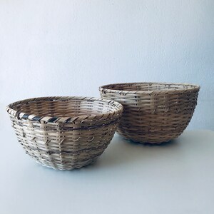Puede incluir: Dos cestas tejidas de materiales naturales. Las cestas son redondas y tienen un color marrón claro con acentos marrones más oscuros. La cesta más grande está detrás de la más pequeña.