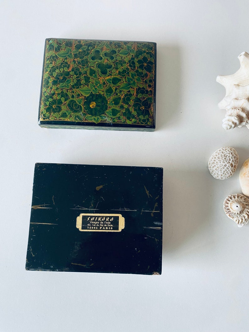 K&ouml;nnte beinhalten: Eine schwarze Box mit einem gr&uuml;nen und goldenen Blumendesign auf der Oberseite. Die Unterseite der Box tr&auml;gt ein Etikett mit der Aufschrift "Shikara, Vases de l'Inde, 40 rue du Faubourg St. Denis, 75010 Paris".