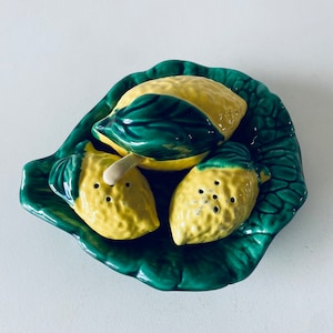 Puede incluir: Un juego de tres saleros y pimenteros de cerámica amarilla con forma de limón sobre una bandeja verde con forma de hoja.