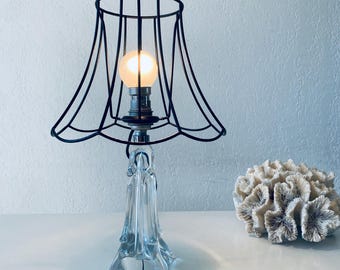 May include: A table lamp with a clear glass base and a black wire cage shade. The lamp is turned on and the light bulb is glowing.
