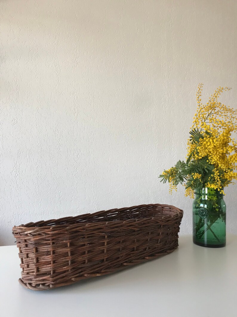 Puede incluir: Una cesta de mimbre larga y ovalada de color marr&oacute;n natural. La cesta est&aacute; sobre una superficie blanca. Un jarr&oacute;n con flores amarillas est&aacute; en el fondo.