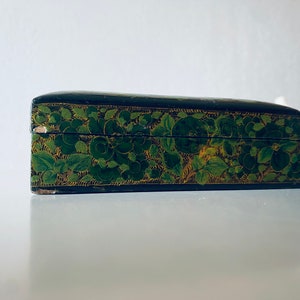 K&ouml;nnte beinhalten: Eine schwarze Holzkiste mit einem gr&uuml;nen und goldenen Blumendesign. Die Kiste ist geschlossen und der Deckel ist leicht ge&ouml;ffnet.