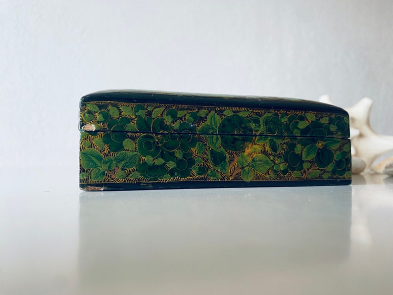 K&ouml;nnte beinhalten: Eine schwarze Holzkiste mit einem gr&uuml;nen und goldenen Blumendesign. Die Kiste ist geschlossen und der Deckel ist leicht ge&ouml;ffnet.
