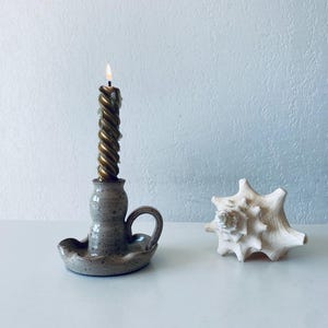 Könnte beinhalten: Ein Kerzenhalter aus Keramik mit einem gesprenkelten braunen und weißen Überzug. Der Halter hat eine gedrehte goldene Kerze im Inneren. Eine weiße Muschel befindet sich auf der rechten Seite des Bildes.