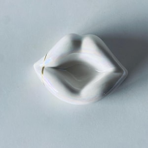 May include: A white ceramic lip-shaped dish with iridescent finish and a gold accent. The dish is a decorative item, possibly for jewelry or small items. The design is minimalist and modern, with a glossy surface.