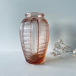 Könnte beinhalten: Eine rosa Glasvase mit geripptem Design und zwei Griffen. Die Vase ist leer und steht auf einer weißen Oberfläche.