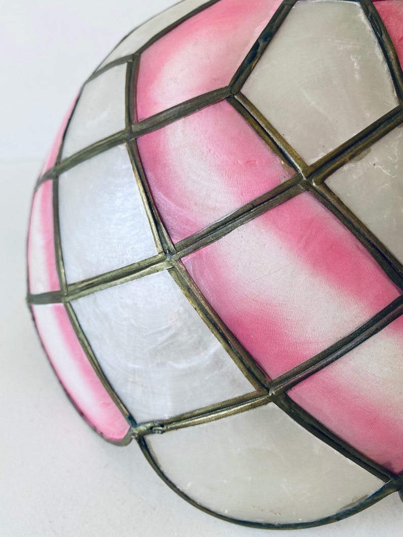 Puede incluir: Una pantalla de l&aacute;mpara de vidrio coloreado rosa y blanco con un patr&oacute;n geom&eacute;trico. La pantalla est&aacute; hecha de paneles de vidrio que se mantienen unidos por un marco de metal.