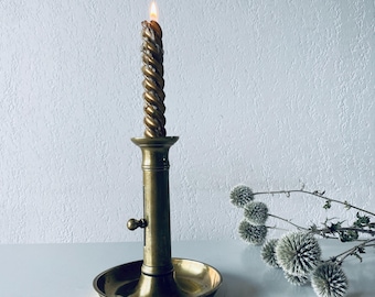 May include: A gold brass candlestick with a twisted candle burning inside. The candlestick has a round base and a small knob on the side.