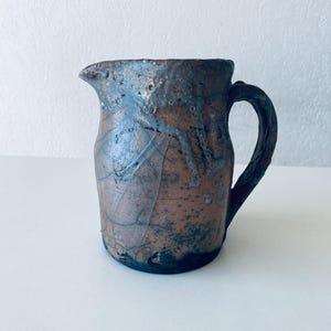 Puede incluir: Una pequeña jarra de cerámica marrón con un esmalte craquelado y un acabado moteado azul oscuro y gris. La jarra tiene un cuerpo redondeado y un asa.