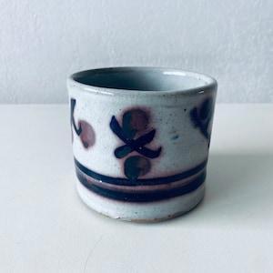 Peut inclure: Une tasse en céramique blanche avec un motif floral violet et bleu. La tasse a un glaçage moucheté et une forme légèrement arrondie.