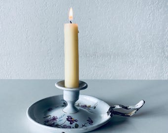 May include: A lit, cream-colored candle sits in a white metal candlestick holder. The holder has a round base with a floral design in purple and green. A handle extends from the side. The candle is burning with a small flame.