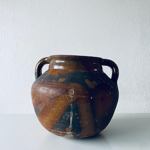 Peut inclure: Un grand vase en céramique marron antique avec deux anses. Le vase a un corps arrondi avec un bord et des anses brun foncé. Le corps présente un mélange de couleurs marron, noir et beige, avec une surface texturée. Le vase est probablement utilisé à des fins décoratives.