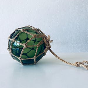 Peut inclure: Un flotteur en verre vert, enfermé dans un filet tissé, avec une corde attachée. Le flotteur est rond et a une surface réfléchissante. La corde est beige et nouée à l'extrémité. Le fond est blanc.