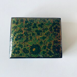 K&ouml;nnte beinhalten: Eine kleine, quadratische Schachtel mit einem gr&uuml;nen und goldenen Blumendesign. Die Schachtel ist aus Holz und hat einen Deckel mit Scharnier.