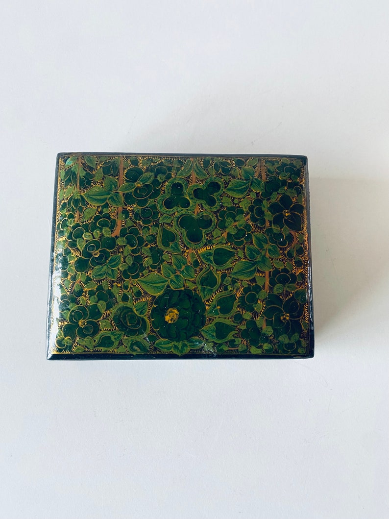 K&ouml;nnte beinhalten: Eine kleine, quadratische Schachtel mit einem gr&uuml;nen und goldenen Blumendesign. Die Schachtel ist aus Holz und hat einen Deckel mit Scharnier.