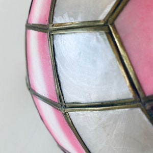 Puede incluir: Una pantalla de l&aacute;mpara geom&eacute;trica rosa y blanca con un marco de lat&oacute;n. La pantalla est&aacute; hecha de un material transl&uacute;cido y tiene un aspecto delicado y femenino.