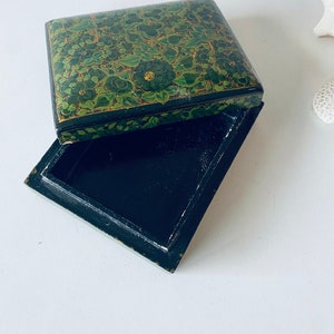 K&ouml;nnte beinhalten: Eine schwarze Holzkiste mit einem gr&uuml;nen und goldenen Blumendesign auf dem Deckel. Die Kiste ist ge&ouml;ffnet und das Innere ist leer.
