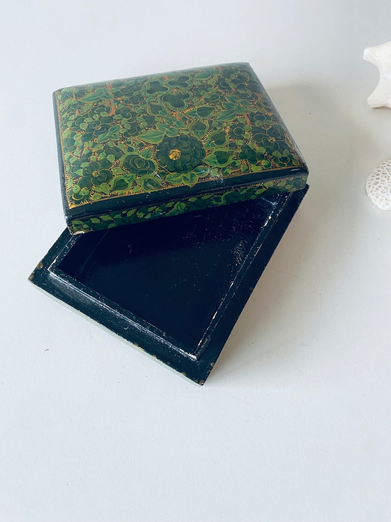 K&ouml;nnte beinhalten: Eine schwarze Holzkiste mit einem gr&uuml;nen und goldenen Blumendesign auf dem Deckel. Die Kiste ist ge&ouml;ffnet und das Innere ist leer.