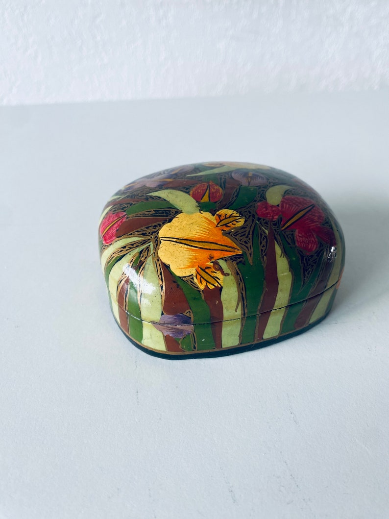 Może przedstawiać: Małe, owalne pudełko z kwiatowym wzorem. Pudełko jest pomalowane zielonymi, brązowymi, ż&oacute;łtymi, czerwonymi i fioletowymi kwiatami. Pudełko jest wykonane z papier-mache.