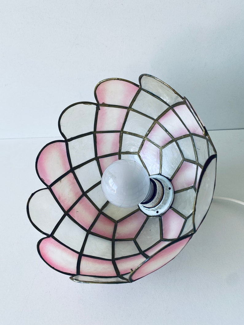 Puede incluir: Una pantalla de l&aacute;mpara rosa y blanca con un patr&oacute;n geom&eacute;trico. La pantalla est&aacute; hecha de un material delgado y transl&uacute;cido y tiene un borde festoneado.