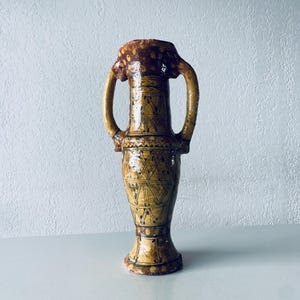 May include: A brown ceramic vase with two handles and a geometric pattern. The vase has a light brown glaze with a darker brown pattern.