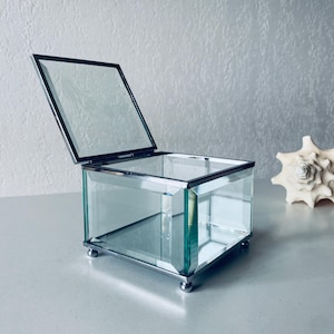 Op de afbeelding: Een kleine, transparante glazen doos met een gespiegelde binnenkant en zilveren rand. De doos heeft een scharnierend deksel en staat op vier kleine voetjes.