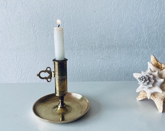 May include: A brass candlestick with a lit white candle. The candleholder has a round base and a decorative handle. A seashell is in the background. The candle is lit with a small flame.