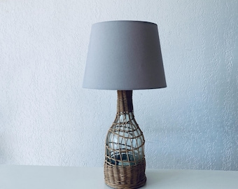 May include: A decorative table lamp with a gray lampshade. The base is a clear glass bottle encased in woven wicker. The lamp is on a white surface against a white textured wall.