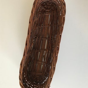 Puede incluir: Una cesta de mimbre marr&oacute;n de forma ovalada alargada. La cesta est&aacute; tejida con finas tiras de madera y tiene un acabado liso y natural.