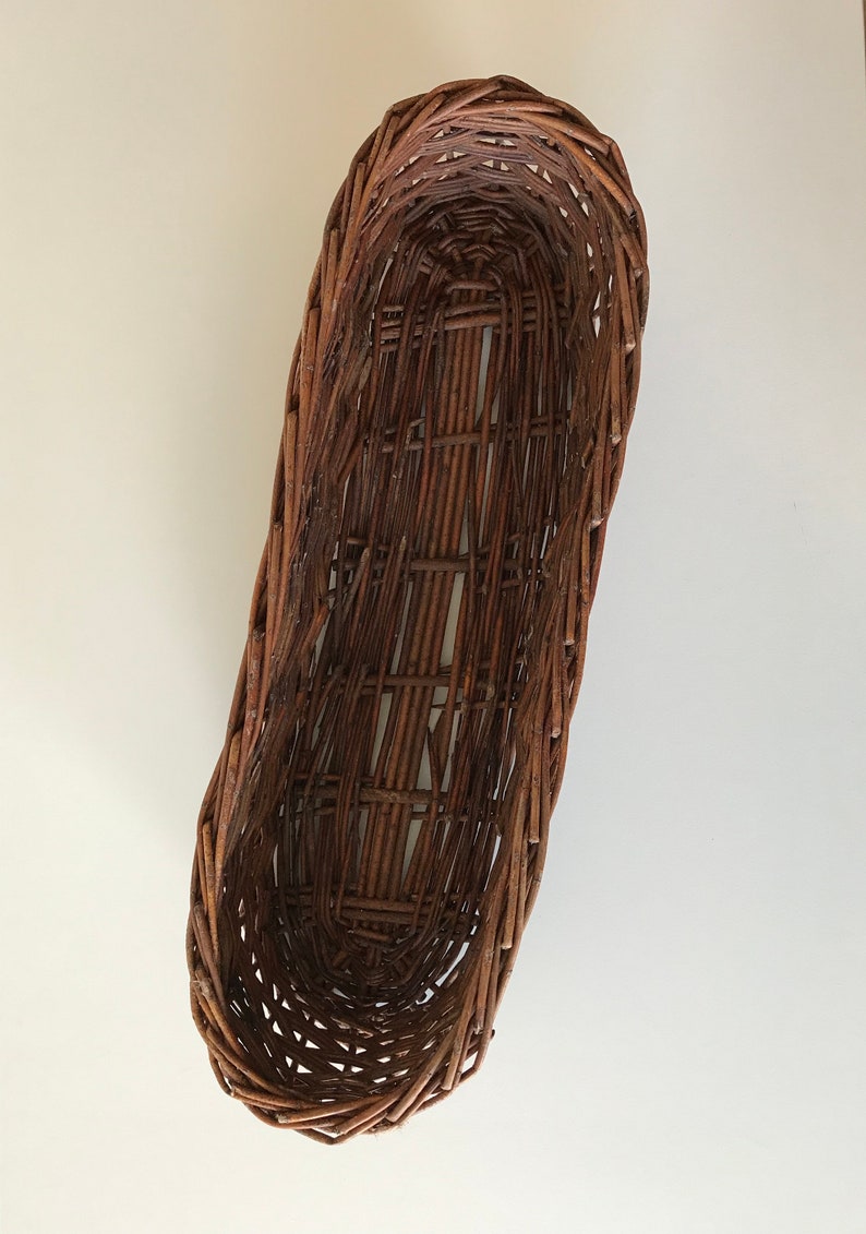 Puede incluir: Una cesta de mimbre marr&oacute;n de forma ovalada alargada. La cesta est&aacute; tejida con finas tiras de madera y tiene un acabado liso y natural.