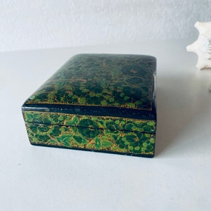 K&ouml;nnte beinhalten: Eine kleine, rechteckige Schachtel mit schwarzem Boden und gr&uuml;nem Blumendesign. Die Schachtel ist mit filigranen Details bemalt und hat einen goldenen Rand.