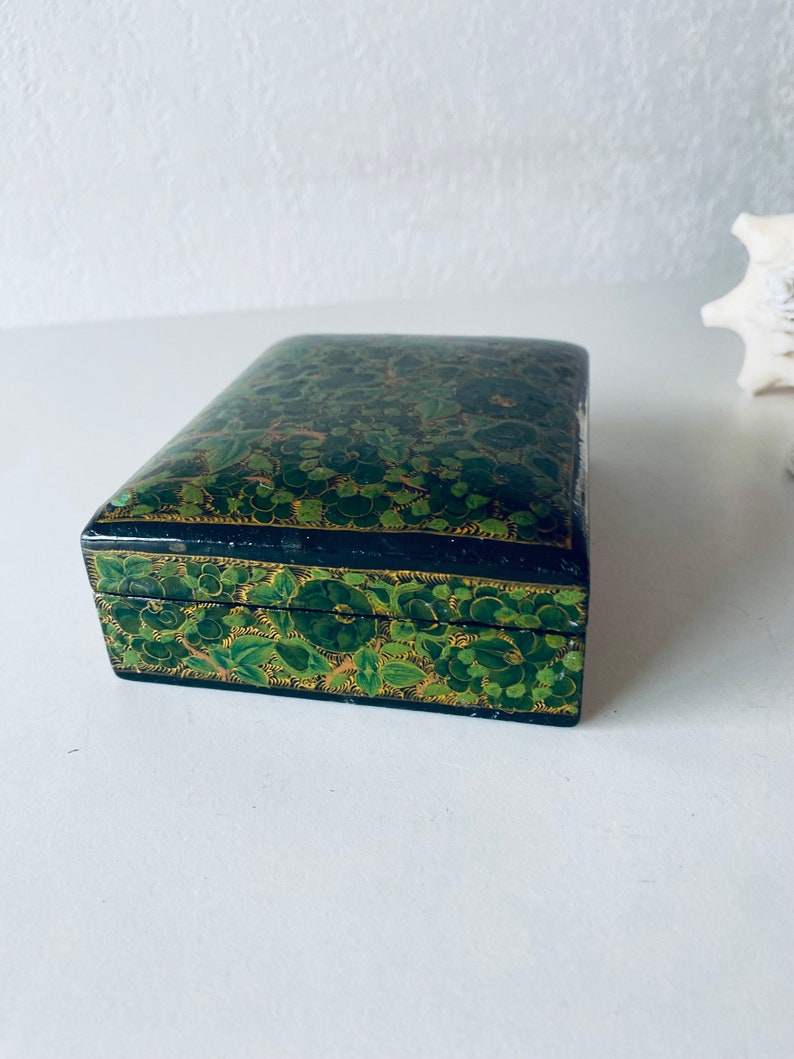 K&ouml;nnte beinhalten: Eine kleine, rechteckige Schachtel mit schwarzem Boden und gr&uuml;nem Blumendesign. Die Schachtel ist mit filigranen Details bemalt und hat einen goldenen Rand.