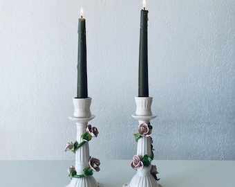 May include: Two white ceramic candle holders with floral details. Each holder features a green vine with pink roses and a black candle.
