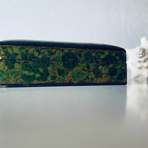 K&ouml;nnte beinhalten: Eine schwarze Holzkiste mit einem gr&uuml;nen und goldenen Blumendesign. Die Kiste ist geschlossen und steht auf einer wei&szlig;en Oberfl&auml;che.